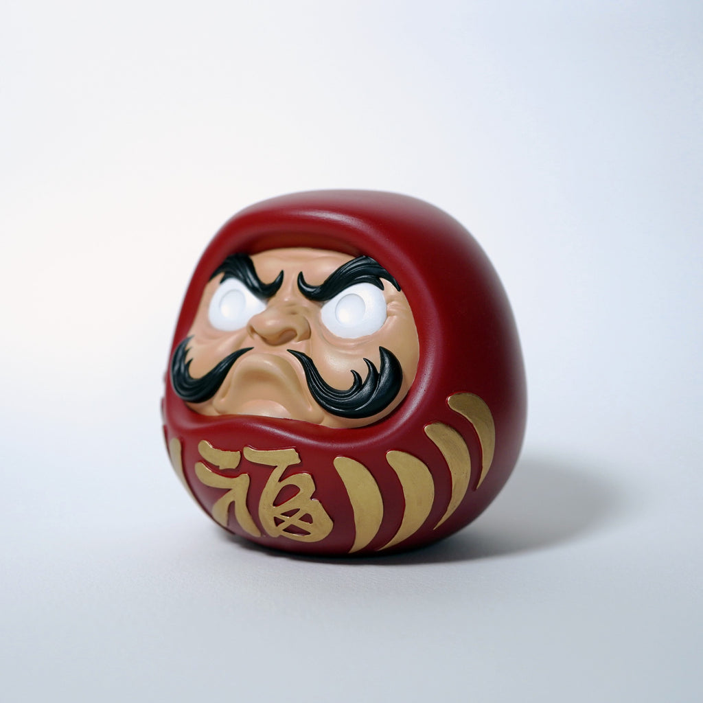 Red Japanese Daruma doll with white eyes and gold kanji, front angled view