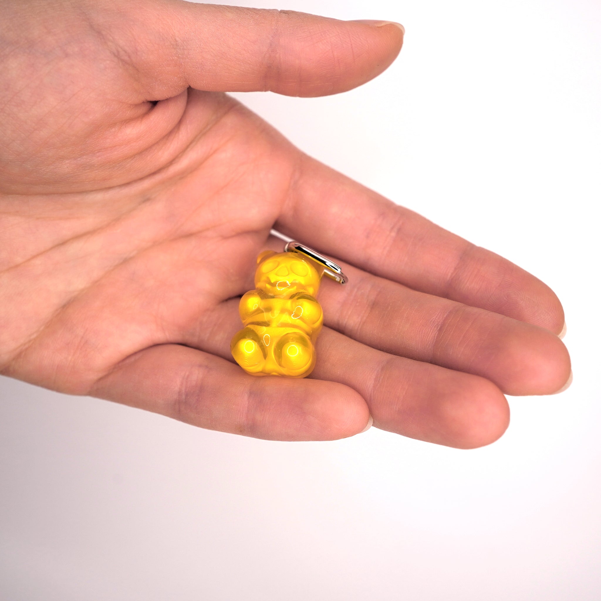 May include: A small, translucent, golden-yellow gummy bear keychain. The bear-shaped charm is attached to a silver-colored keyring. The image is set against a white background.