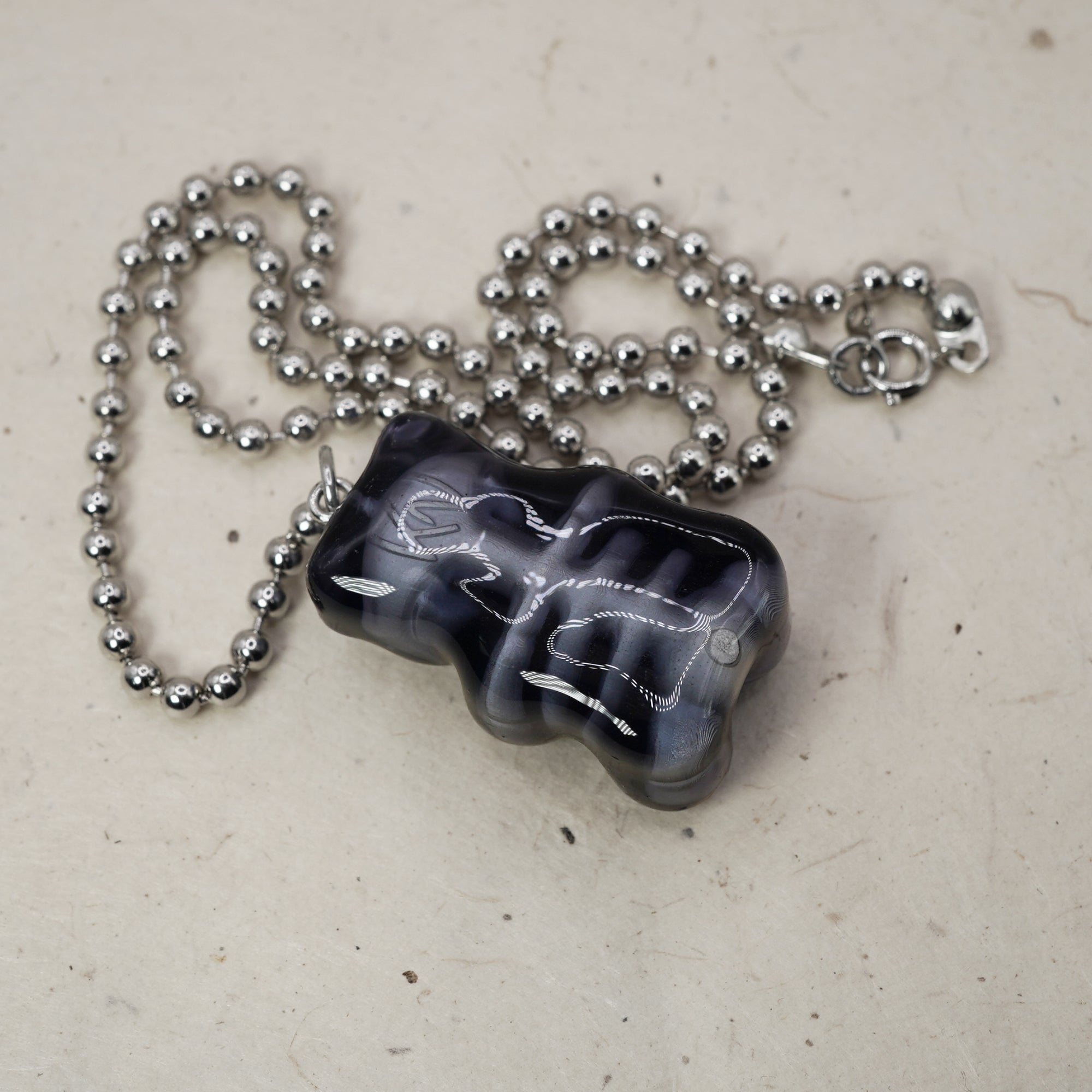 May include: A dark gray and white marbled pendant necklace. The pendant is a rounded, abstract shape with white veining. The necklace chain is made of small, silver-colored ball beads and has a clasp.