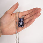 Black X-ray gummy bear pendant shown in hand for scale, glossy resin finish with visible white skeleton, silver ball chain necklace.