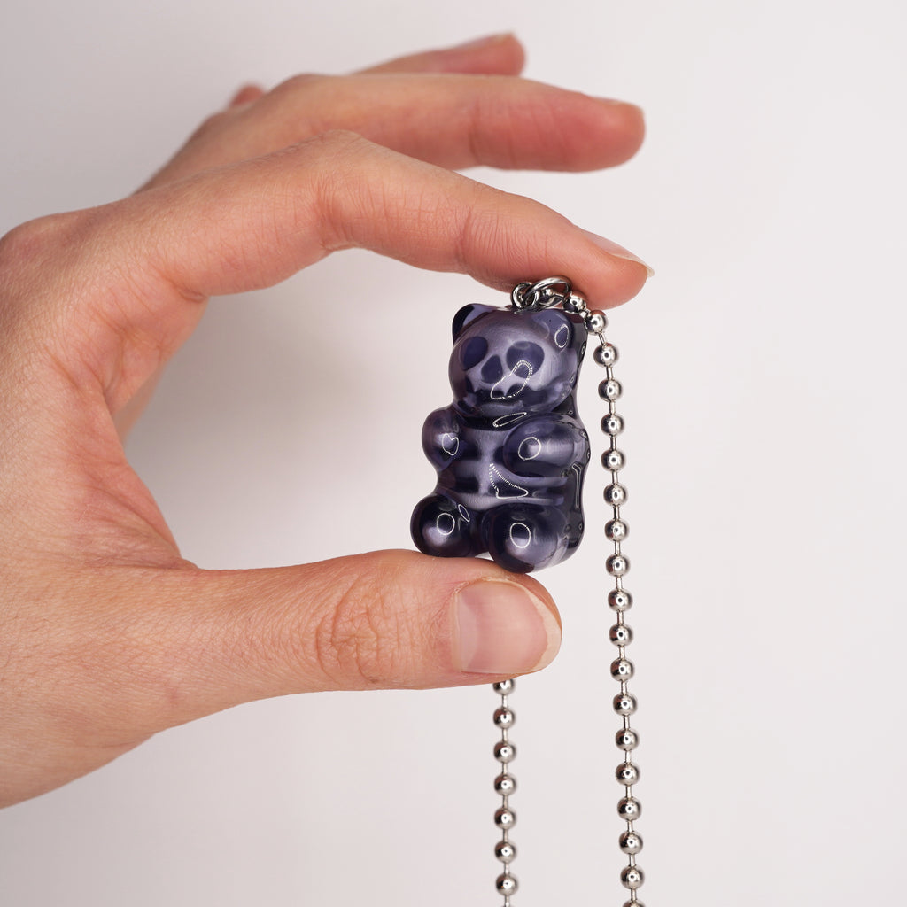 May include: A dark purple gummy bear pendant on a silver ball chain. The bear has a detailed design with lighter purple highlights. The pendant is held up against a white background.