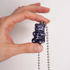 May include: A dark purple gummy bear pendant on a silver ball chain. The bear has a detailed design with lighter purple highlights. The pendant is held up against a white background.