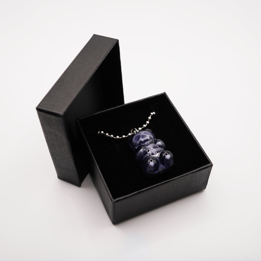 A dark blue gummy bear pendant necklace displayed in a black gift box. The pendant is made of a translucent material and hangs from a silver chain. The box is open, revealing the necklace against a black velvet interior.