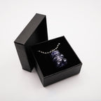 A dark blue gummy bear pendant necklace displayed in a black gift box. The pendant is made of a translucent material and hangs from a silver chain. The box is open, revealing the necklace against a black velvet interior.