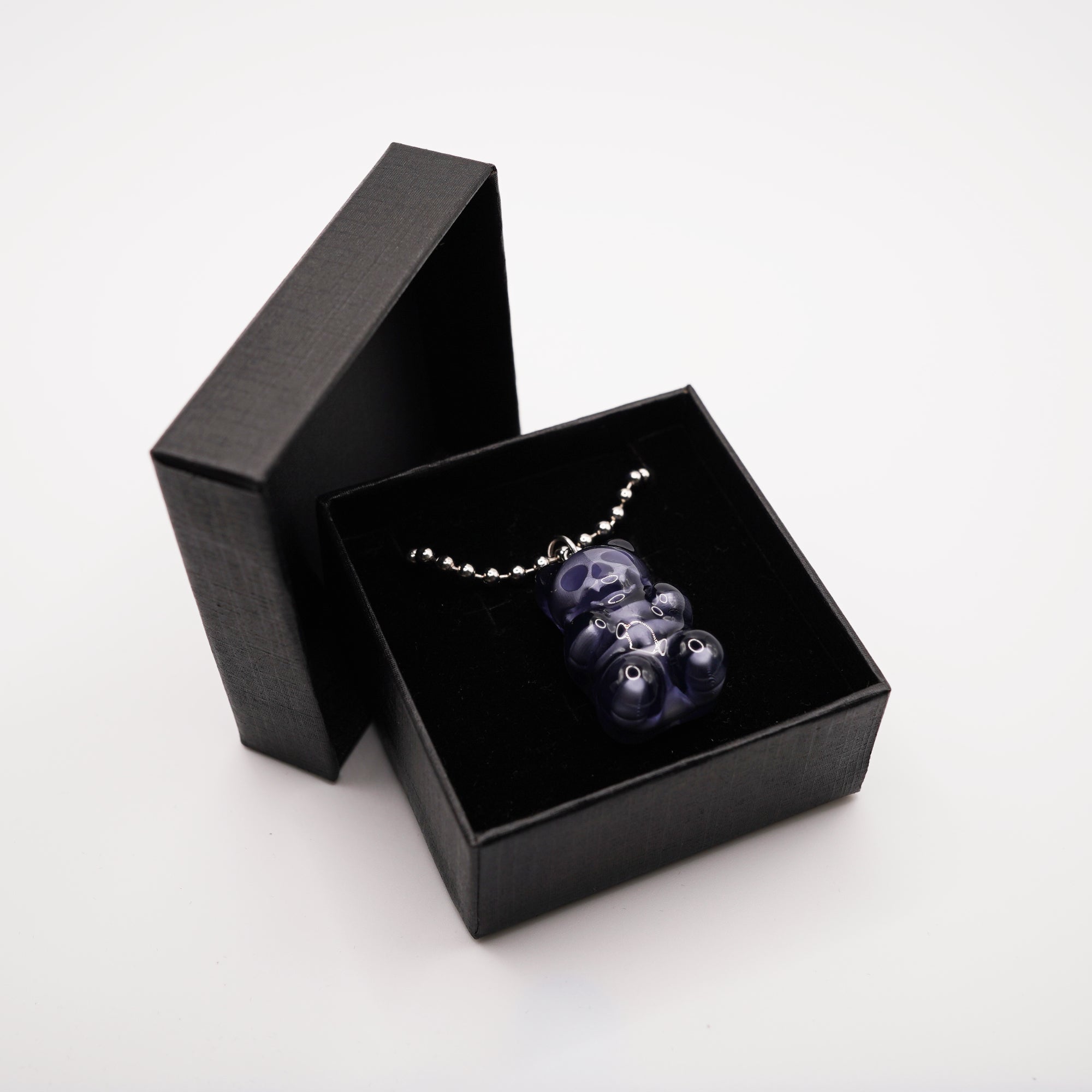 A dark blue gummy bear pendant necklace displayed in a black gift box. The pendant is made of a translucent material and hangs from a silver chain. The box is open, revealing the necklace against a black velvet interior.