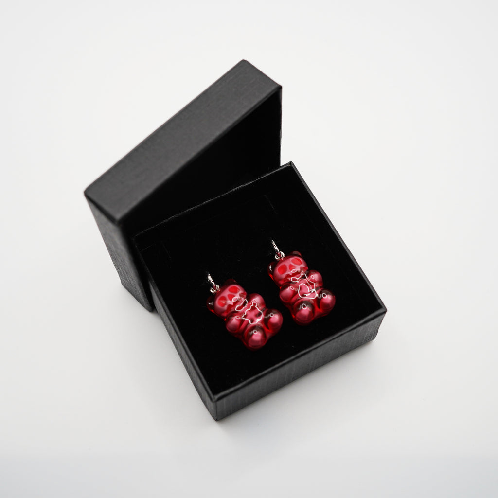May include: A pair of red gummy bear earrings with silver hooks, presented in a black gift box. The earrings have a glossy finish and are detailed with white accents. The box is open, revealing the earrings against a black interior.