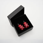 May include: A pair of red gummy bear earrings with silver hooks, presented in a black gift box. The earrings have a glossy finish and are detailed with white accents. The box is open, revealing the earrings against a black interior.
