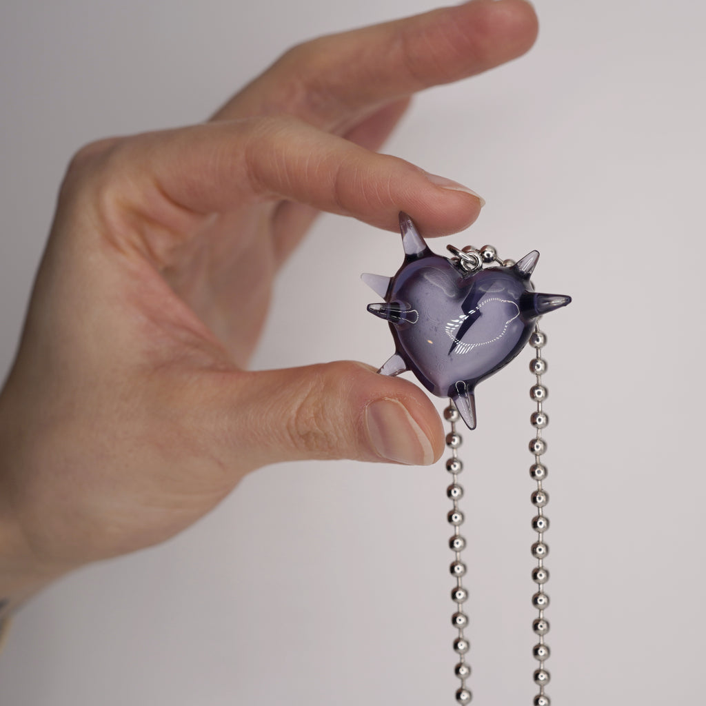 A purple heart-shaped pendant necklace with a lightning bolt design. The pendant has spiky protrusions and hangs from a silver ball chain. The necklace is held up against a white background.